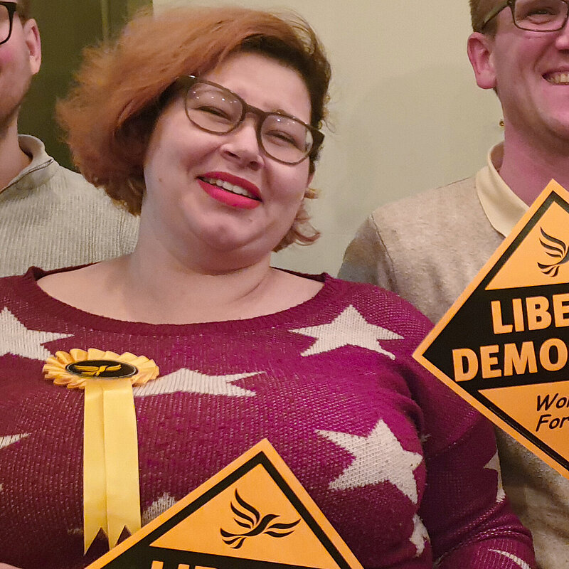 Photo of Sophie Aldridge, Diversity & Inclusivity Officer for the Hertsmere Lib Dems, smiling while carrying a "Liberal Democrats Winning Here" poster.