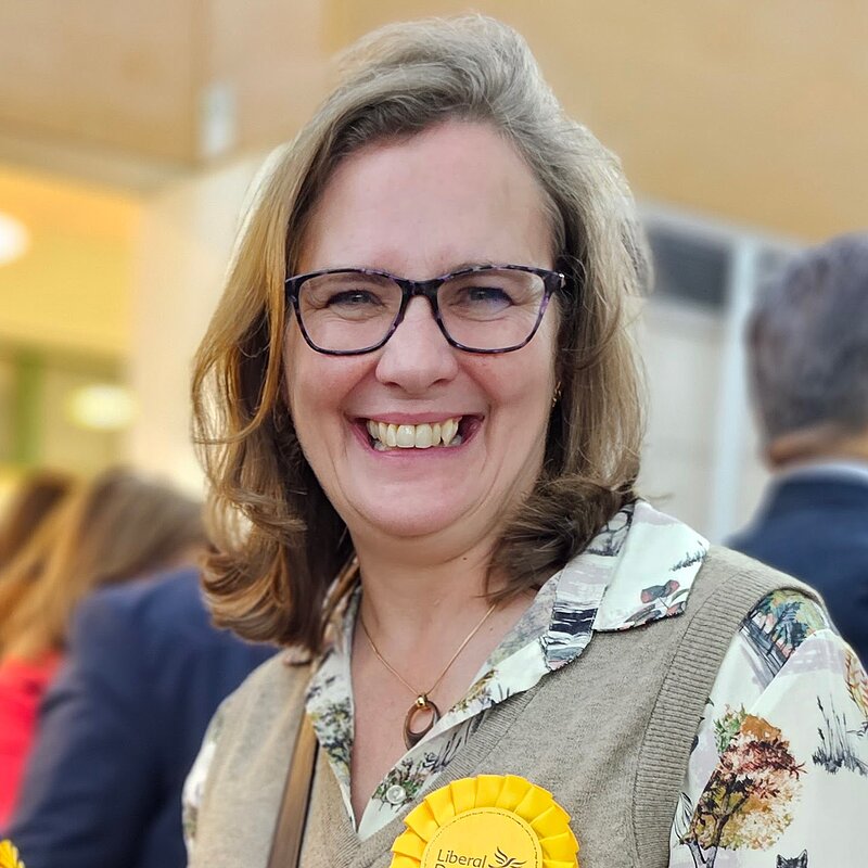 Photo of Susan Barrett, Secretary for the Hertsmere Lib Dems, smiling while wearing a Liberal Democrats rosette.